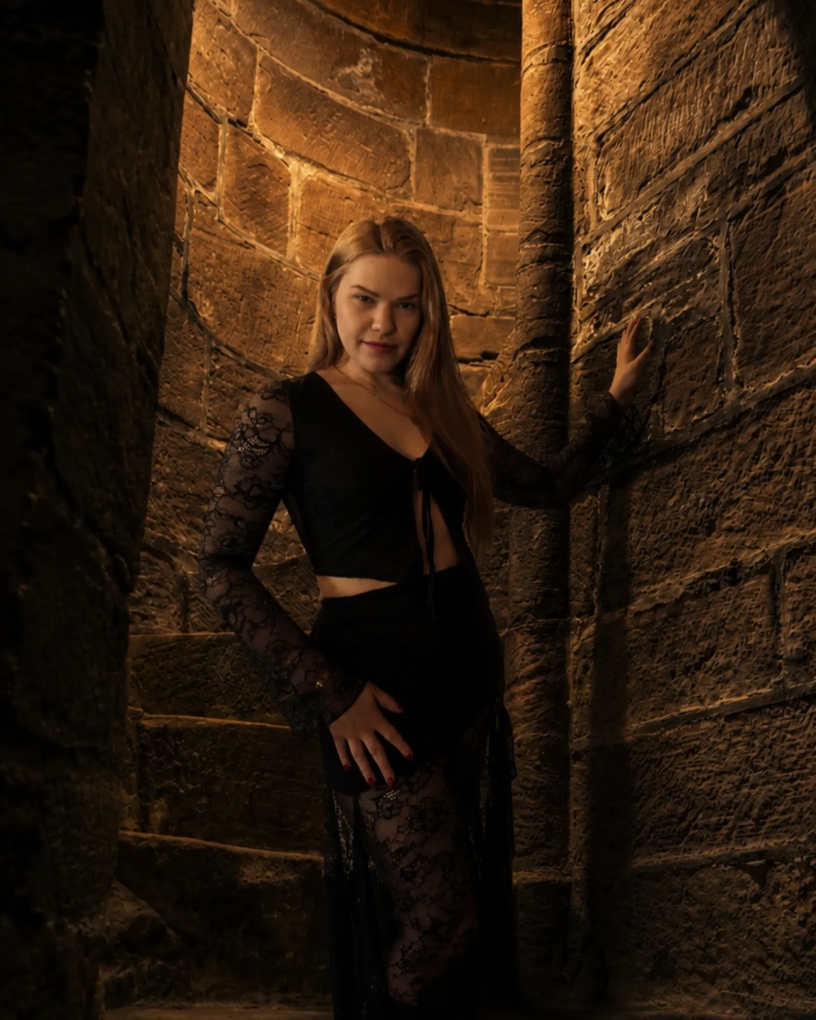 Toronto dominatrix Princess Stella standing in a narrow stone corridor lit by warm amber light.